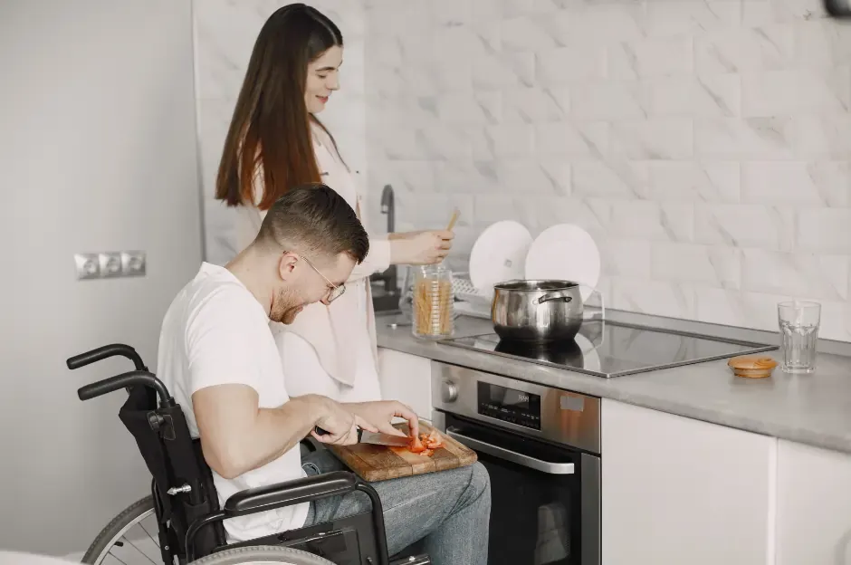 The Impact of Proper Home Modifications on Quality of Life - Home modifications reduce hours of care provided by 42% per week. disabled man in wheelchair and woman standing in kitchen at modified benchtop that is lower for wheelchair user to be able to cook for themself.