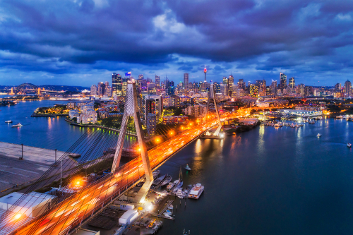 The Anzac Bridge, captured at dusk, its cables and pylons dramatically lit against the fading light. The bridge serves as a vital link and a poignant memorial, with the Australian and New Zealand flags flying high, symbolizing the enduring spirit of camaraderie and sacrifice.