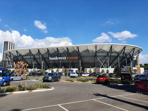 The vibrant atmosphere of Bankwest Stadium during a night game, with the stands filled with cheering fans, the field brightly lit, and the action underway. The architectural design of the stadium, with its close seating arrangement, ensures an intimate and intense spectator experience, capturing the spirit of community and sporting excellence in Parramatta. 