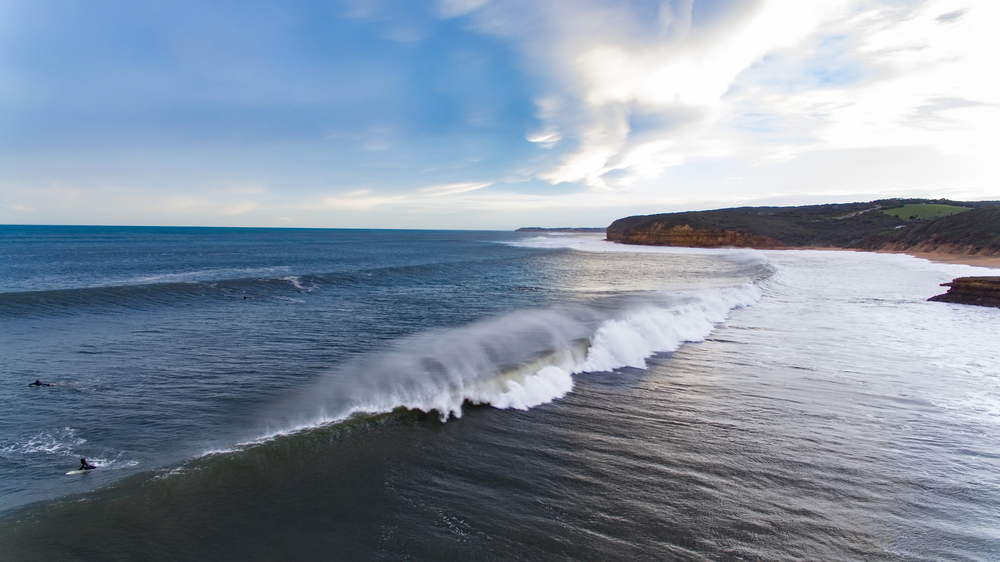A vibrant image of surfers riding the renowned swells of Bells Beach during a surfing competition. Spectators line the beach, watching as surfers tackle some of the best waves Australia has to offer. The backdrop features the rugged coastline and the surf community's enthusiastic spirit, showcasing Bells Beach as a premier destination for international surf events and everyday surf enthusiasts alike.