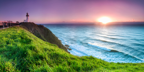The idyllic beachfront of Byron Bay, capturing its famous lighthouse, pristine beaches, and surfers riding the waves, all set against a backdrop of a vibrant town known for its arts, music, and relaxed coastal culture.