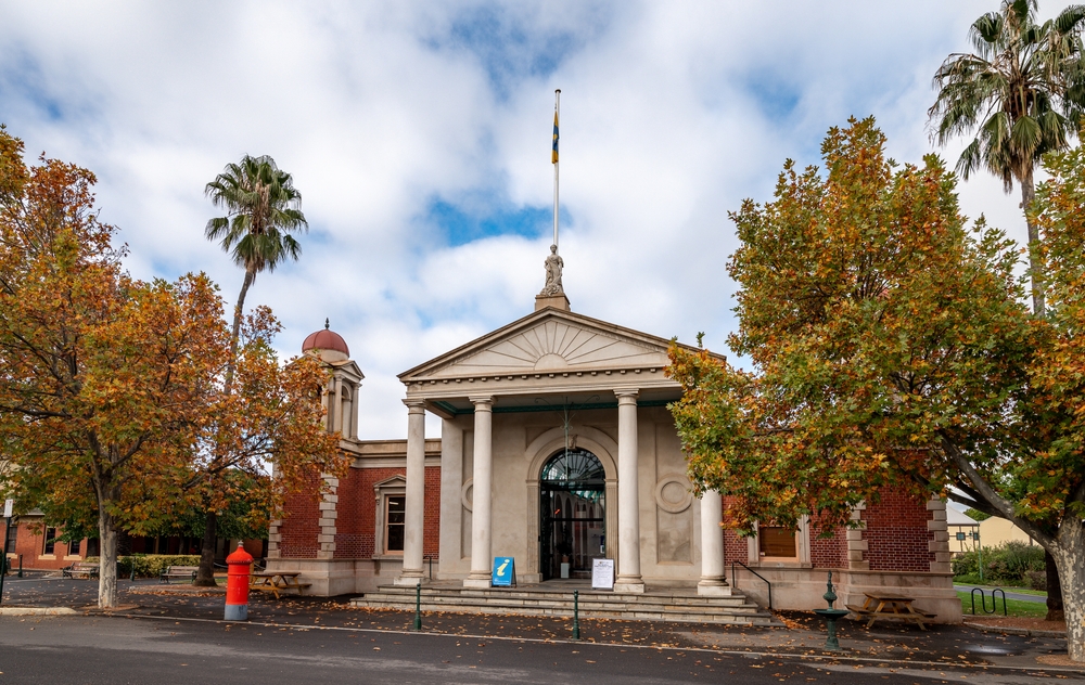 An image showcasing the exterior of the Castlemaine Art Museum, highlighting its elegant Art Deco facade which blends seamlessly with modern architectural elements. The building stands prominently on a sunny day, its clean lines and geometric forms drawing the eye and inviting visitors inside. The museum's setting amid well-maintained gardens and its position as a cultural landmark in Castlemaine make it a focal point for art lovers and history enthusiasts alike.