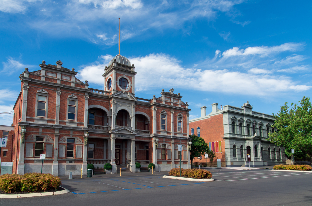 A picturesque view of the Castlemaine Town Hall, captured in the golden light of late afternoon, emphasizes its grand Victorian architecture. The Town Hall, with its intricate brickwork, large arched windows, and a classic clock tower, serves as a stunning example of the region's rich heritage. This image portrays the building as a bustling center of community life, where history and modern-day events coalesce.