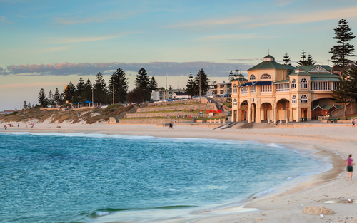 This panoramic view captures the essence of Cottesloe Beach with its clear turquoise waters and golden sands, stretching invitingly under a vibrant blue sky. In the background, the iconic Indiana Tea House stands as a prominent feature, embodying the beach's historic charm. The scene is alive with people enjoying various activities, from swimming to surfing, against the backdrop of a stunning sunset. Sculptures dot the shoreline, adding an artistic flair to the natural beauty, illustrating the beach's role as an outdoor art gallery during the Sculpture by the Sea exhibition.