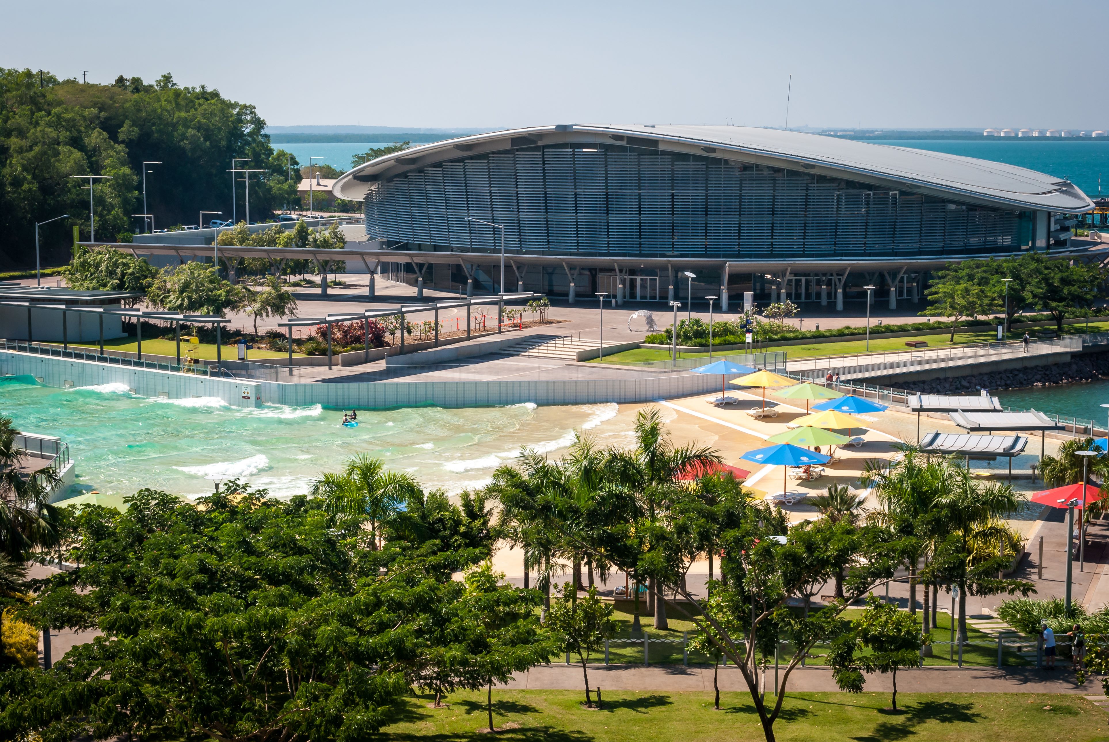  A photo of the Darwin Waterfront, with people enjoying the wave pool and lagoon, surrounded by palm trees and modern buildings. 