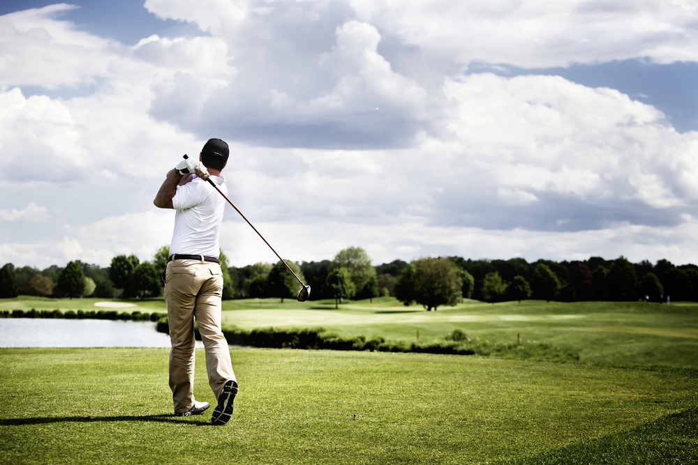 An image showcasing the lush, expansive fairways of Eynesbury Golf Course, set against a backdrop of natural bushland under a clear blue sky. Golfers can be seen in mid-swing, focusing intently on their game, while native birds and wildlife add a vibrant touch of nature to the scene. The meticulously maintained greenery and strategically placed bunkers highlight the course's reputation for quality and challenge. This picturesque setting not only attracts golf enthusiasts but also those who appreciate the serene beauty and historical charm of Eynesbury.