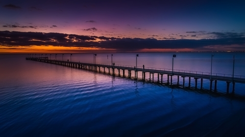 unset view of Frankston Pier extending into Port Phillip Bay, featuring renovated walking paths and seating areas, with visitors enjoying the serene waterfront atmosphere.