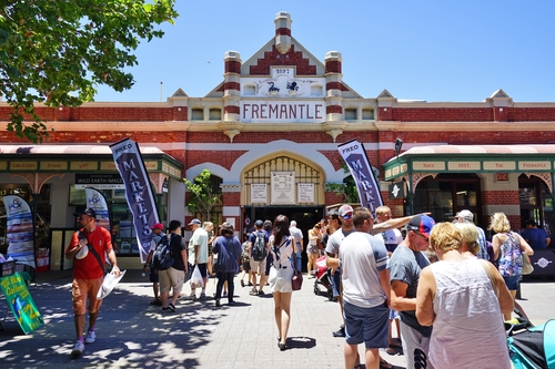 A vibrant and bustling scene at the Fremantle Markets, showcasing a wide array of stalls selling fresh produce, artisan goods, and gourmet foods. The heritage architecture of the market building adds a historic charm, with colorful signage and lively crowds of shoppers exploring the diverse offerings.