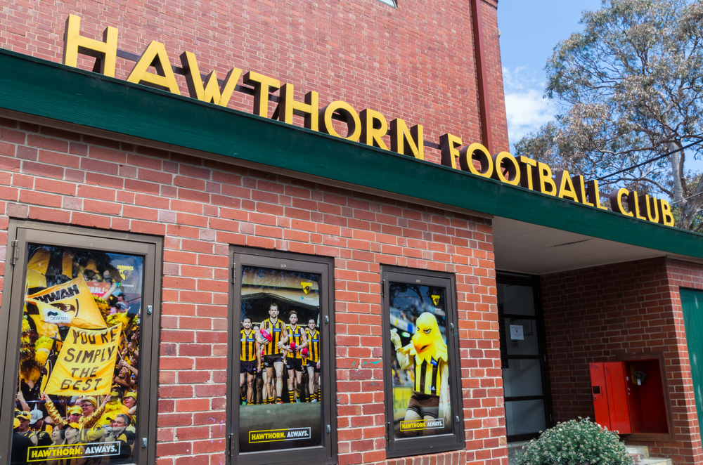 An image depicting the exterior of Glenferrie Oval, highlighting its classic facade and entrance gate adorned with historical signage. The architecture reflects the traditional style of early 20th-century sports stadiums, with its red brick walls and green iron gates that evoke a nostalgic sporting heritage. This view captures the essence of the venue's long-standing connection to Australian Rules football and its iconic status in the community of Hawthorn.