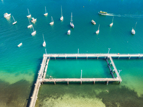 The serene setting of Gunnamatta Bay, with its clear blue waters and boats gently bobbing at the marina. Families and friends gather on the shores and jetties, enjoying the tranquil atmosphere and the leisurely pace of life that defines the Sutherland Shire's waterside communities.