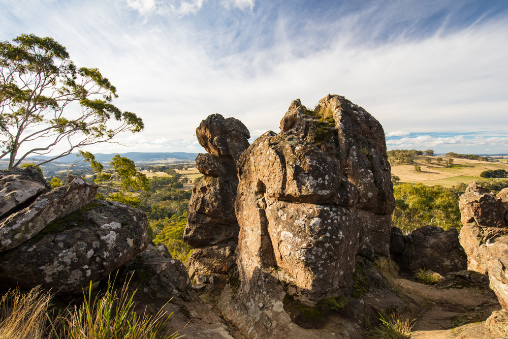 An image capturing the mystical presence of Hanging Rock at sunset, with the rock formations dramatically silhouetted against a vibrant sky. This natural volcanic feature rises starkly from the surrounding green landscape, drawing visitors with its eerie beauty and the mysterious legends encapsulated by the famous novel and film. The trails and picnic areas are subtly visible, inviting exploration and leisure in this enchanting setting.