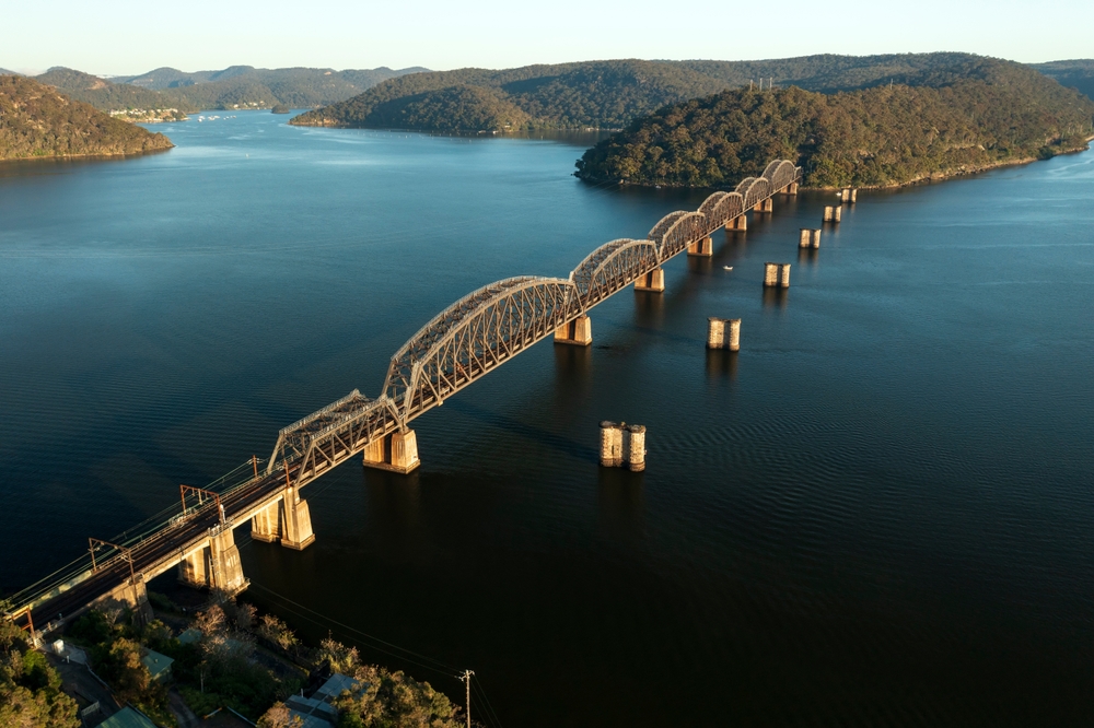 A striking image of the Hawkesbury River Railroad Bridge. The bridge spans the wide river, connecting the rocky riverbanks, and is framed by the natural bushland of the surrounding national parks.