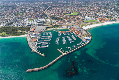 Capture the vibrant atmosphere of Hillarys Boat Harbour with its colorful array of shops, restaurants, and boats docked in the marina, against the backdrop of a bustling crowd enjoying the waterfront views and activities.