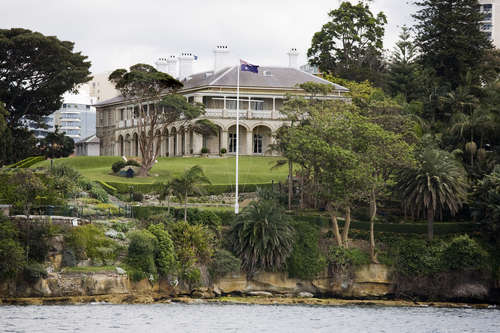 Kirribilli House, seen from a vantage point that showcases its grandeur and the lush gardens that surround it. The Sydney Harbour and Opera House form a stunning backdrop, highlighting the residence's prestigious location and architectural elegance.
