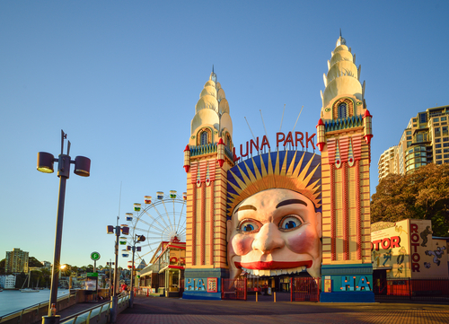 The vibrant entrance to Luna Park, with its iconic smiling face welcoming visitors into a world of amusement and laughter. Inside, the park buzzes with excitement, from the colourful carousel to the thrilling Ferris wheel, against the backdrop of Sydney Harbour, encapsulating the timeless appeal of this historic amusement park.