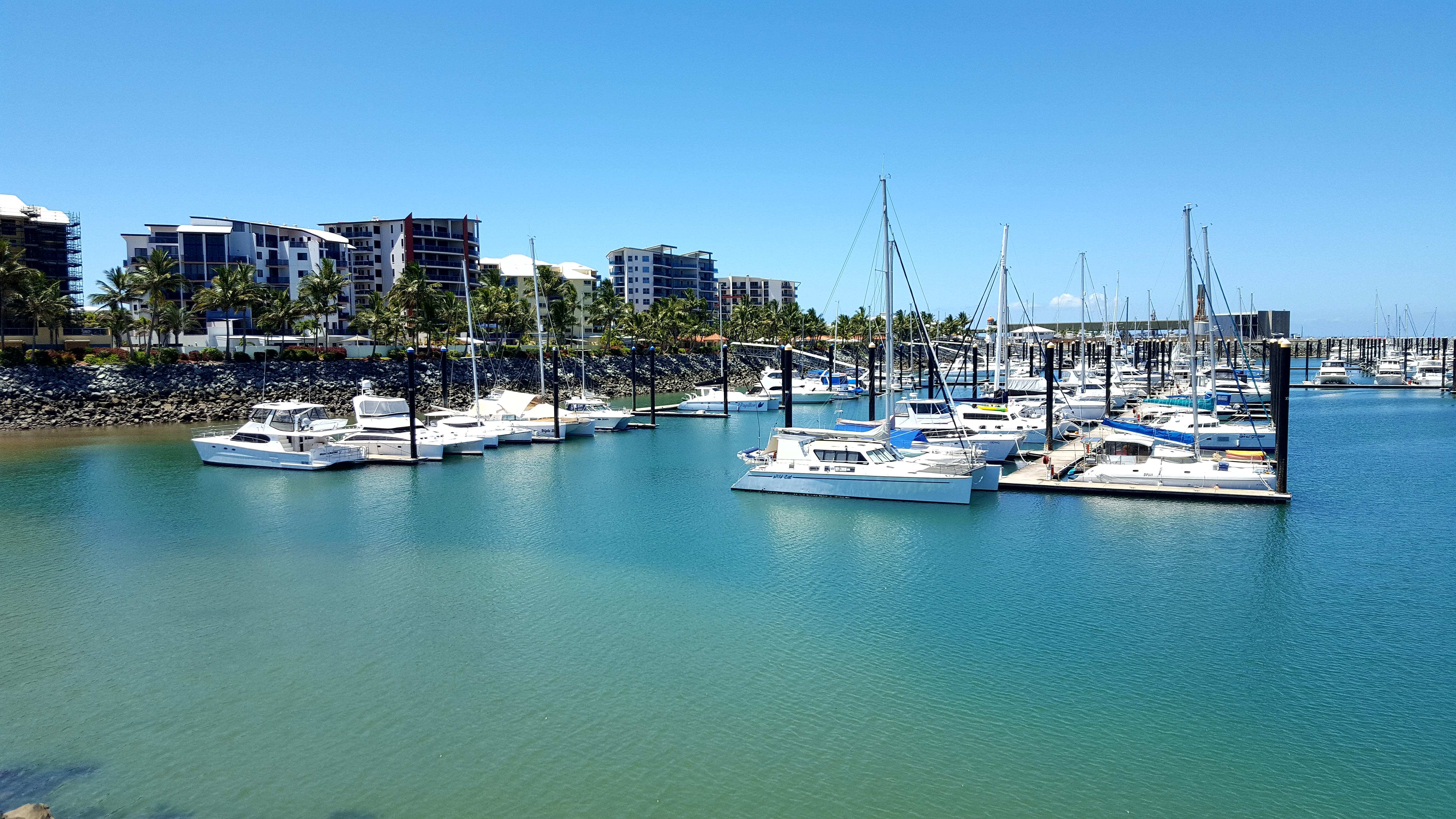 A scenic image of Mackay Marina, with boats docked along the marina, waterfront cafes, and a view of the cityscape. 
