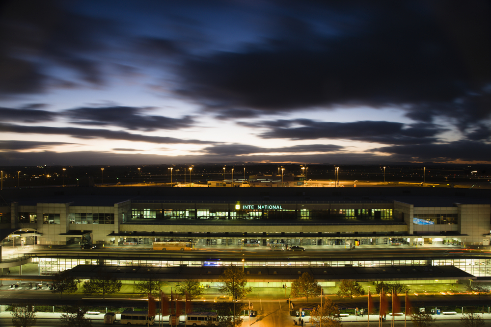 An image capturing the exterior of Melbourne Airport, highlighting its expansive entrance and modern facade. The airport's grand scale is evident, with wide access roads bustling with a variety of vehicles — from taxis and buses to private cars — all converging at the drop-off and pick-up zones. The terminal's large glass windows reflect the sky, giving a glimpse of the airport's busy atmosphere and its role as a major international gateway. This image portrays Melbourne Airport as a vital infrastructure hub, bustling with activity and connecting Melbourne to the world.