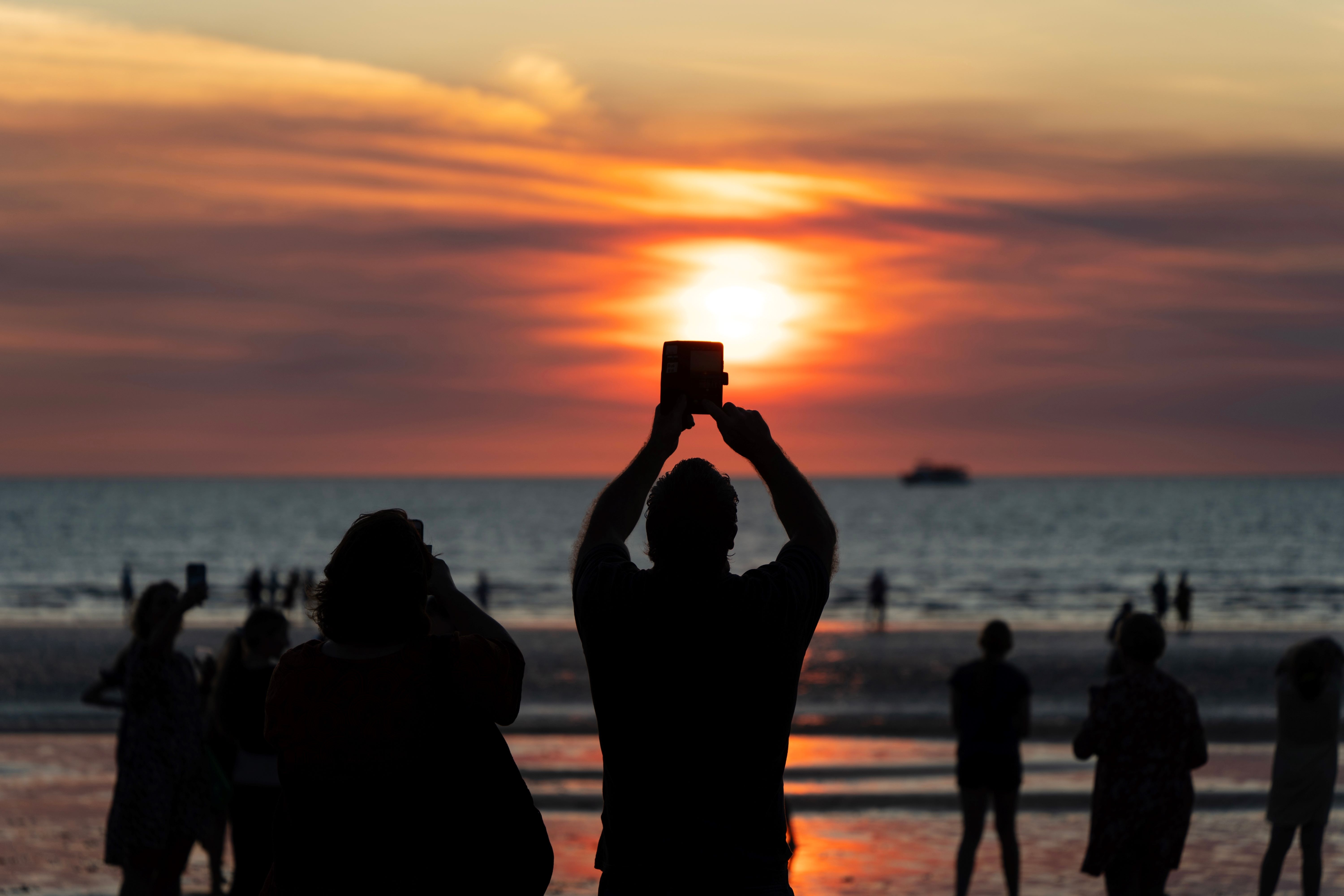 A panoramic view of the Mindil Beach Sunset Market, with colorful stalls lining the beach, people browsing, and the sun setting in the distance. 