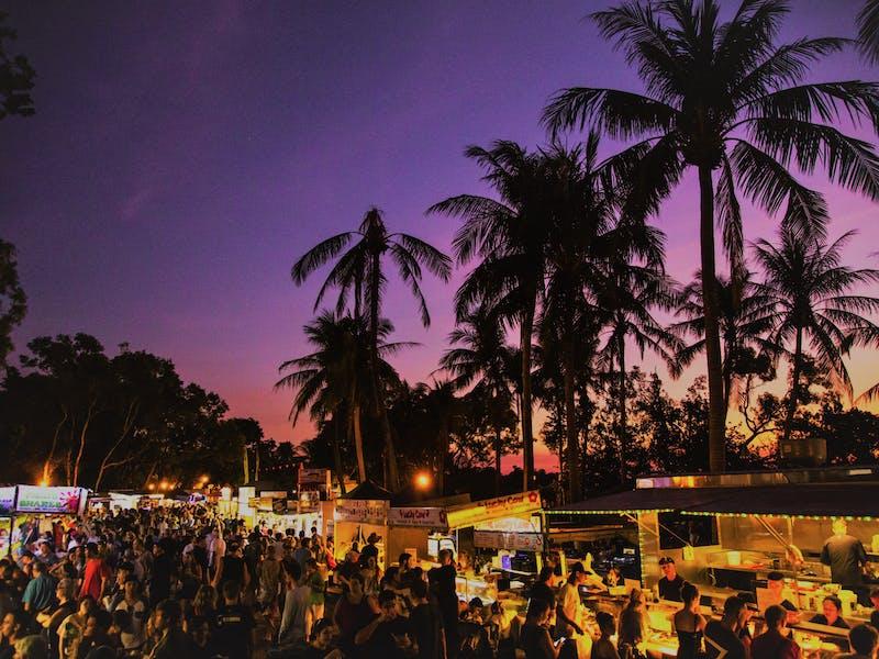 An atmospheric snapshot of Mindil Beach Sunset Market at dusk, with vibrant stalls illuminated by the setting sun, people enjoying the beachfront atmosphere, and the sky ablaze with fiery hues, creating a magical ambiance for all to enjoy. 