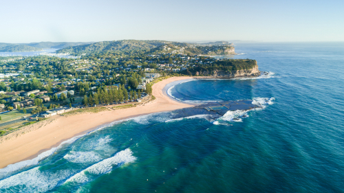 A tranquil morning at Mona Vale Beach, with soft sunlight illuminating the surf and sand. Surfers dot the waves in search of the perfect ride, while early walkers enjoy the peaceful ambiance along the shoreline. The rock pool, a distinctive feature of the beach, offers a calm spot for swimmers against the backdrop of the beach's natural beauty.