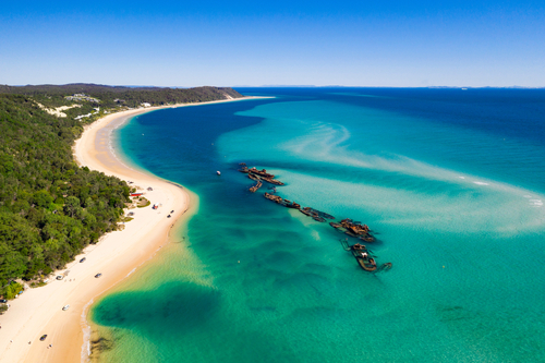 An image capturing the essence of Moreton Island with its expansive, sandy beaches and towering sand dunes under a clear blue sky. In the foreground, a family enjoys the sun-soaked beach, with a backdrop of the island's unique landscapes, including the famous Tangalooma Wrecks visible in the distance, offering a perfect blend of natural beauty and adventure.