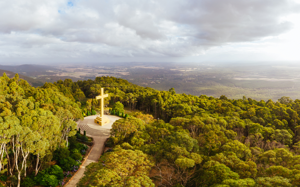 A powerful image of the Mount Macedon Memorial Cross standing solemnly against a sweeping vista of the Macedon Ranges. The cross, a towering structure, is set against a backdrop of dense eucalyptus forest and expansive skies, offering a place of reflection and remembrance. Visitors can be seen gathered around the base, some taking in the panoramic views that stretch out over the region, symbolizing peace and sacrifice.