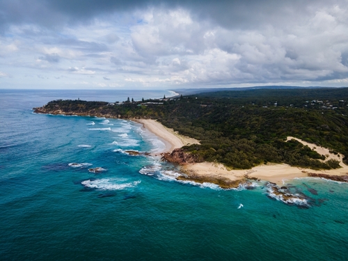 A serene image of North Stradbroke Island, highlighting its pristine Point Lookout with panoramic ocean views where visitors can spot whales breaching the surface. The picture combines the rugged coastal beauty, with waves crashing against rocky cliffs, and the tranquil, clear waters, embodying the island's blend of cultural significance and natural wonders.