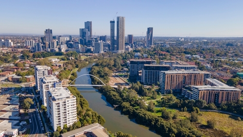 A serene view of Parramatta River at sunset, with the water reflecting the changing colours of the sky and the city's skyline in the distance. People are seen enjoying the riverside, either strolling along the paths, fishing, or simply taking in the picturesque environment, highlighting the river's role as a cherished natural asset in the urban setting. 