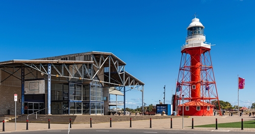 A vibrant image that combines the historic Port Adelaide Lighthouse, standing tall and proud, with the bustling energy of the Fishermen's Wharf Market in the foreground. Shoppers explore stalls filled with antiques, crafts, and local delicacies, under the watchful guide of the lighthouse. The market is alive with the community's spirit, showcasing the rich history and cultural vibrancy of Port Adelaide. This picture perfectly captures the essence of the Western Suburbs as a hub of maritime heritage and lively local commerce.