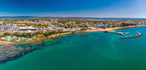 A vibrant snapshot of the Redcliffe Peninsula, showcasing its picturesque seaside promenade lined with blooming trees and bustling cafes. The focal point is the Bee Gees Way, a lively outdoor museum featuring statues and memorabilia of the iconic band, under the warm glow of evening lights, reflecting the area's rich musical heritage and community spirit.