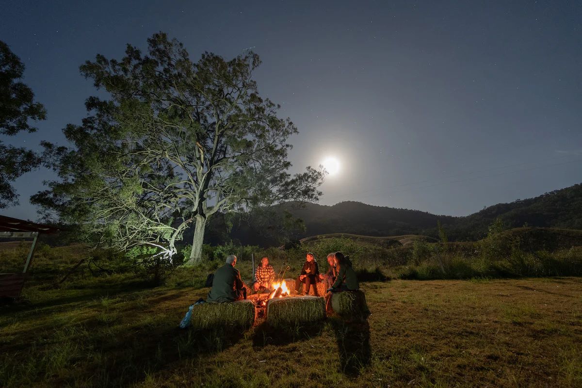 An Indigenous elder sharing Dreaming stories with visitors around a campfire under the night sky.