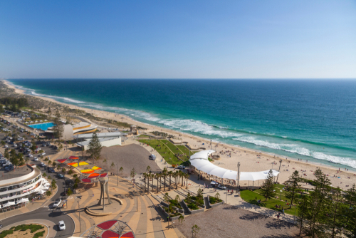 A panoramic view of Scarborough Beach showcases its vibrant atmosphere, with surfers riding the waves, families enjoying the golden sands, and the lively foreshore area bustling with activity. The clear, turquoise waters contrast beautifully with the sunny, blue sky, making it a picturesque destination for beach lovers.