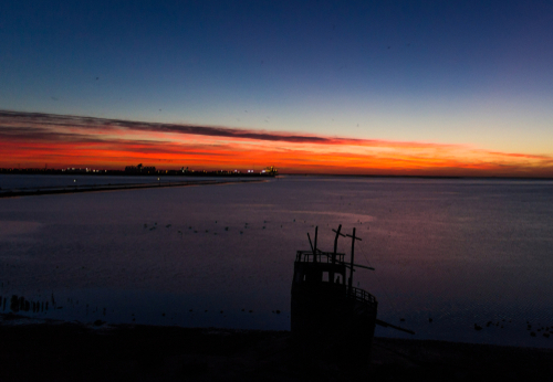 An image captures the striking silhouette of the shipwreck play structure at St Kilda Adventure Playground, set against the vibrant hues of a sunset sky. The playground, transformed into an explorer's paradise, allows children's imaginations to sail the high seas. The fading sunlight casts a golden glow over the scene, highlighting the playground's unique attraction and the beauty of outdoor play in the Northern Suburbs of Adelaide.