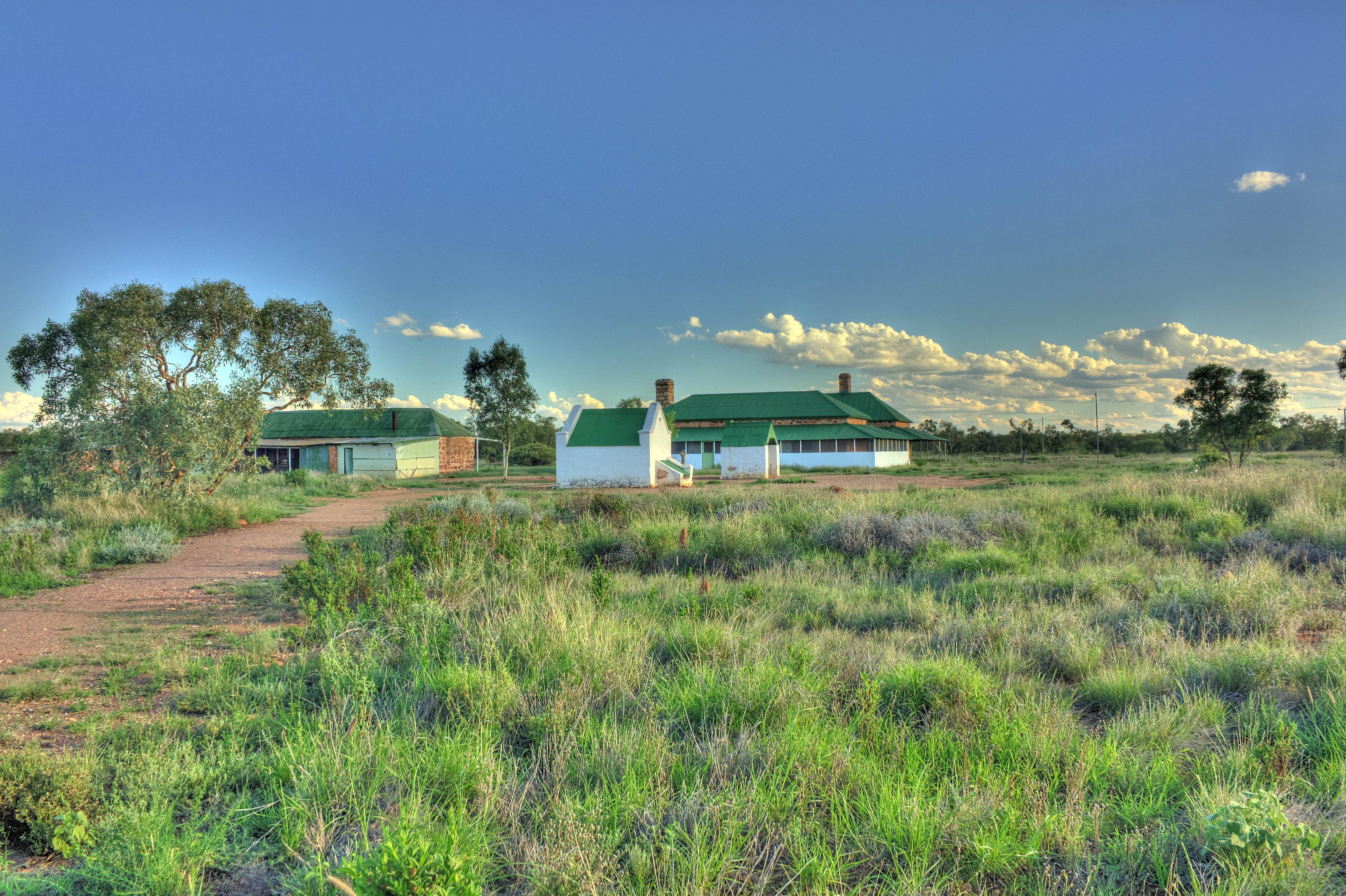A restored telegraph station building with a red corrugated iron roof, set against a backdrop of Australian outback landscape. 