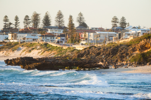 Trigg Beach, captured in the serene light of early morning, reveals its appeal to surfers and nature enthusiasts. The beach is framed by natural bushland, offering scenic walking trails that lead to peaceful picnic spots. In the foreground, consistent waves invite surfers to enjoy the surf, while in the background, families set up for a day by the sea.