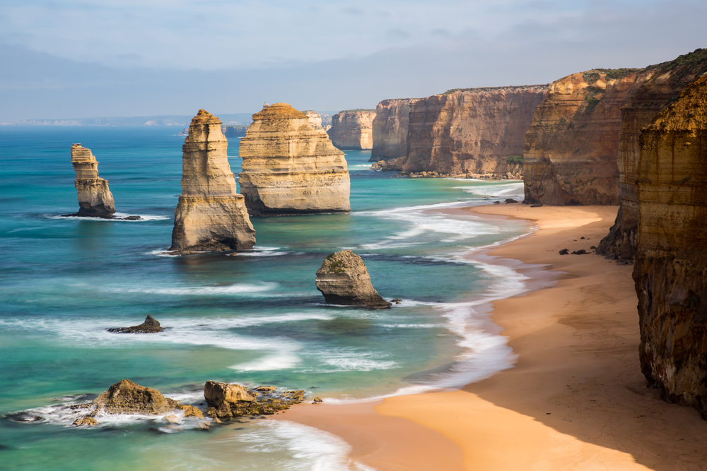 An image capturing the stunning Twelve Apostles at sunset, where the golden light illuminates the massive limestone stacks that rise dramatically from the Southern Ocean. The ocean's powerful waves crash against the base of the cliffs, highlighting the natural erosion process that shaped these famous formations. The viewpoint is filled with awe-struck visitors, emphasizing the global appeal and breathtaking beauty of this natural wonder along the Great Ocean Road.