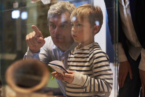 A heartwarming image depicts a child and father exploring the Uleybury School Museum together, their curiosity piqued by the historical artifacts before them. They are seen examining an old-fashioned classroom setup, complete with wooden desks and historical educational tools. The museum's interior, bathed in natural light, creates an intimate setting for this educational journey, underscoring the museum's role in connecting generations through the rich tapestry of local history.