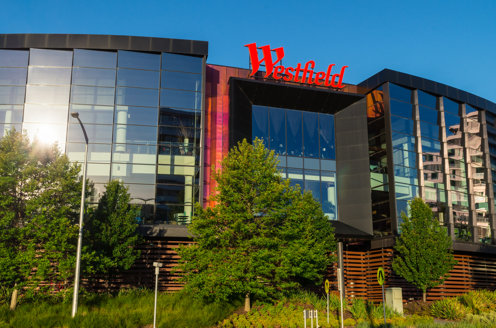 An external view of Westfield Doncaster shows the shopping center's modern and sleek architecture characterized by expansive glass panels and a streamlined, contemporary design. The exterior is bustling with activity, featuring shoppers entering and exiting through the prominently displayed entrances, surrounded by landscaped areas that enhance the building's upscale urban appeal. This image illustrates the mall as a beacon of modern retail and lifestyle within Inner Eastern Melbourne.
