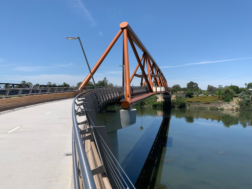 A panoramic view of the Yandhai Nepean Crossing at sunrise, showcasing its elegant design and the breathtaking backdrop of the Blue Mountains. The bridge is alive with early morning joggers and cyclists, symbolizing the community's vibrant spirit and the blend of natural beauty and cultural significance that defines Penrith.
