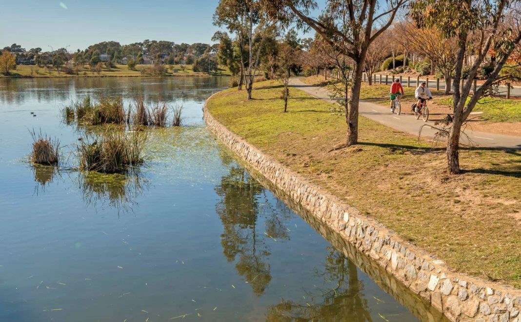 A serene view of Yerrabi Pond, with lush greenery framing the tranquil waters, and a walking path winding along the shoreline under a clear blue sky.  