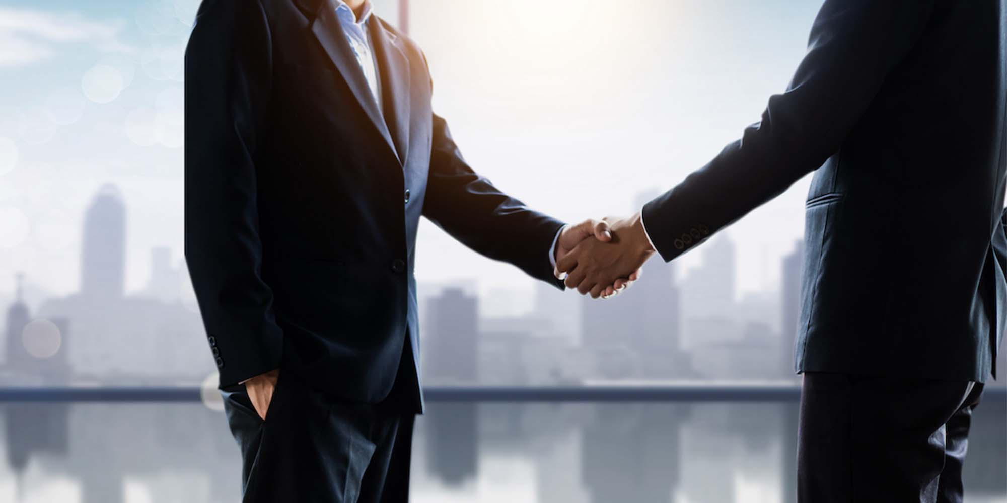 Two business men in suits shaking hands over a city view sky line