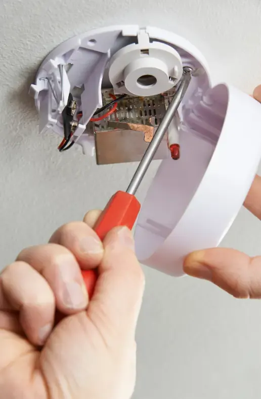 smoke alarm that has been opened by electrician who is holding a screwdriver in the smoke detector to test it and make sure it is working as it should