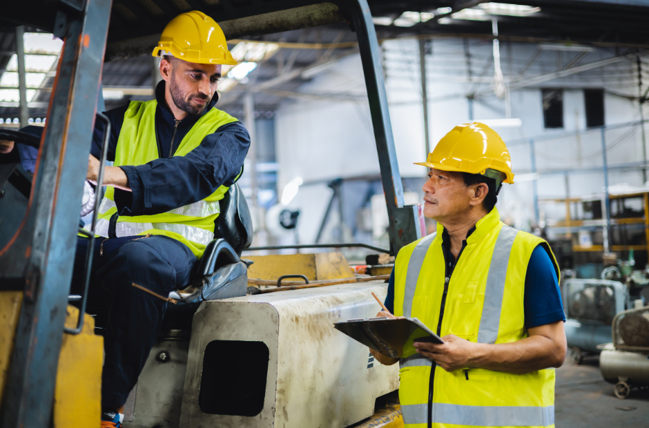 two employees with a checklist for the pre-start safety inspection on a forklift. both are wearing hard hats and fluro vests in a warehouse where forklifts are being used